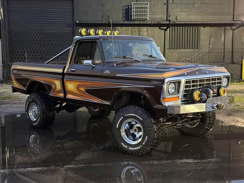 Brownie, a lifted brown 1978 Ford F-150 with a KC light bar and winch bumper, parked on wet asphalt outside a shop at dusk