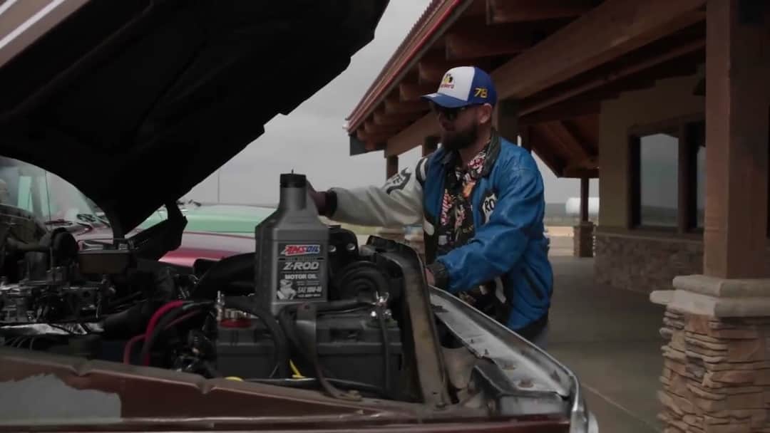Solomon reaching into Brownie's open engine bay under a stone-pillared canopy, a quart bottle of AMSOIL motor oil standing on the engine beside his hand.