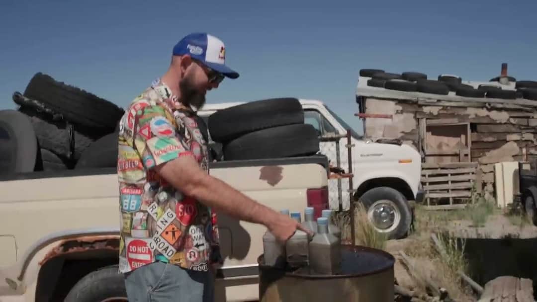 Solomon in a Route 66 patterned shirt pointing at a row of grey quart bottles standing on an oil drum, a white van and stacked tyres behind him in the desert.