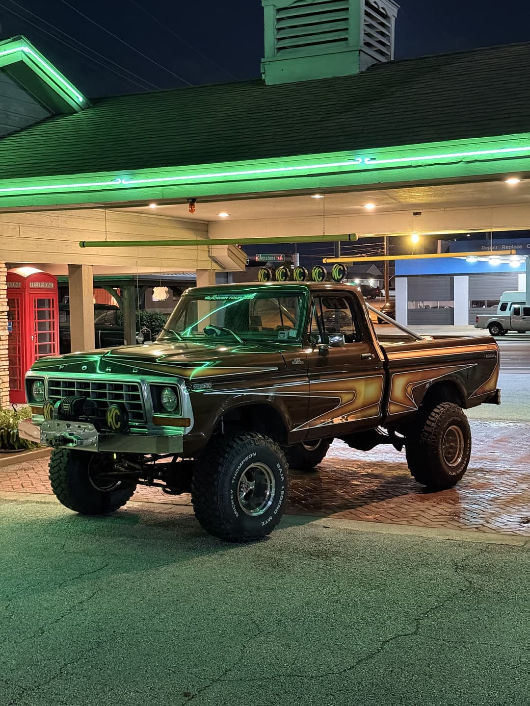 Brownie parked under the canopy of a restored service station at night, green neon running along the eaves and reflecting off the hood, a red telephone box at the left of frame and wet brick paving underfoot.