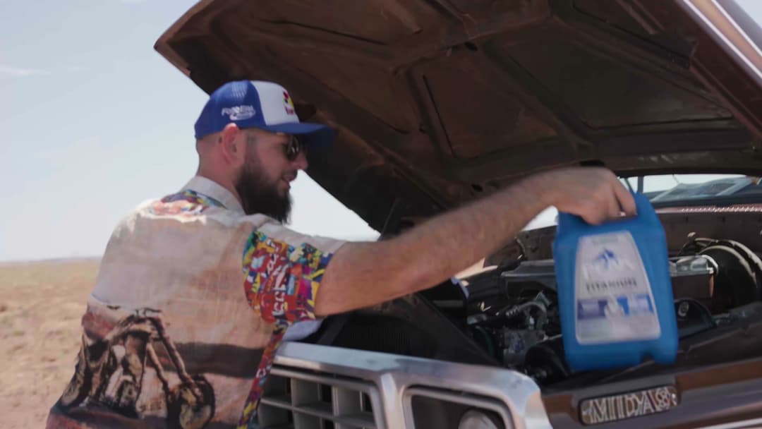 Solomon lifting a blue PEAK coolant jug beside Brownie's open hood on a bright desert roadside, the truck and open landscape filling the frame.