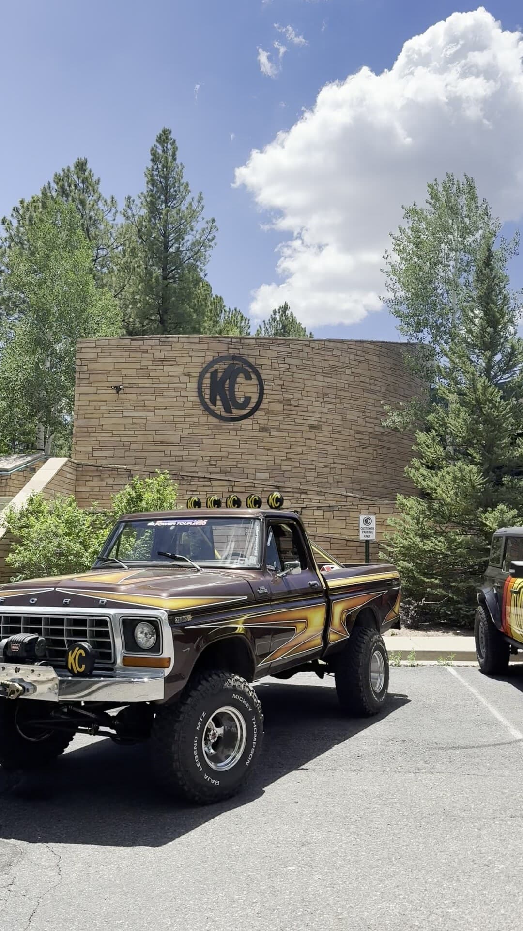 Brownie, a 1978 Ford F-150, parked in front of the KC HiLiTES stone monument in Flagstaff. The KC mark is mounted on the stacked-stone wall above the truck, five KC lights sit on the roll bar and one on the front bumper, with pines and a clouded blue sky behind.