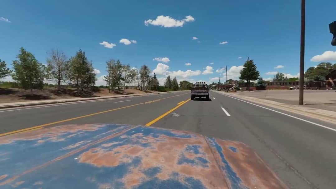 The view forward over a patinated hood on a wide Flagstaff highway, Brownie ahead in the same lane with its roll-bar lights showing against a clouded blue sky.