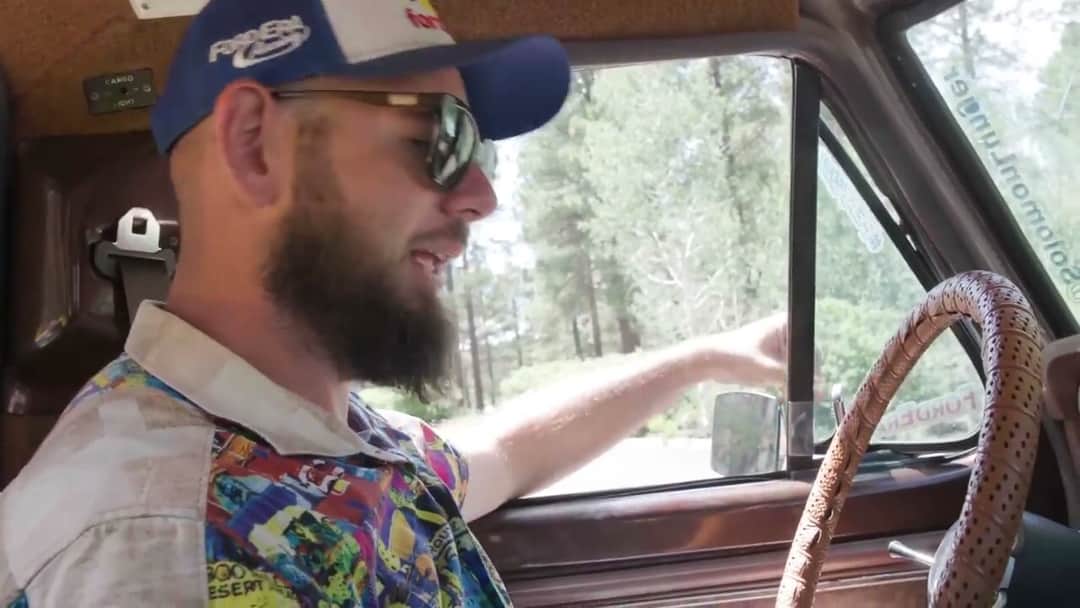 Solomon at the wheel of Brownie in sunglasses and a patterned shirt, one hand on the wooden steering wheel, pines running past the open window.